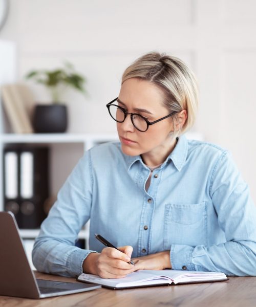 Focused Caucasian woman writing down info during business training or online meeting, participating in web conference from home. Smart female student taking distance training course